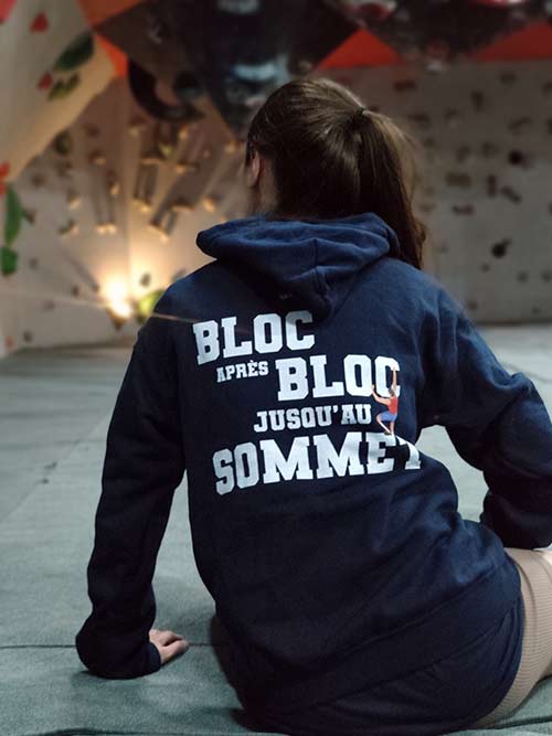 Femme assise de dos dans une salle de bloc portant le sweat à capuche bleu marine Bloc après bloc, jusqu'au sommet