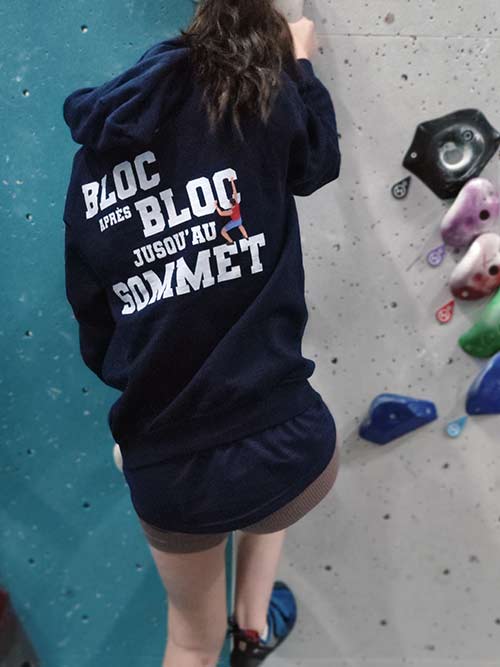 Femme de dos en train de grimper sur un mur de bloc avec le sweat à capuche bleu marine Bloc après bloc, jusqu'au sommet